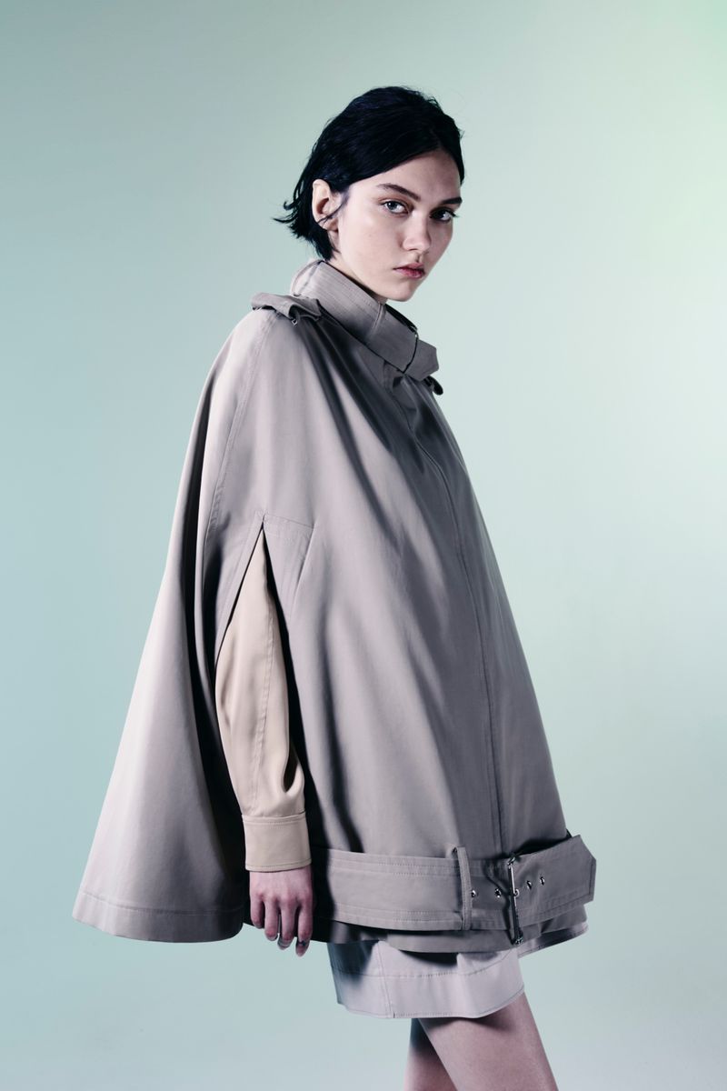 Fashion portrait of a woman wearing a tan cape looking at the camera against a solid background in a studio with diffused light.
