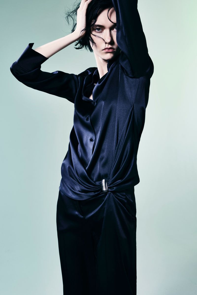 An adult female model poses in a dark blue satin-like blouse and matching pants against a light green background in a studio shot.