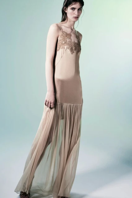 A slender woman poses in a flowing, tan gown with lace detailing, against a light blue background in a fashion editorial style shot.