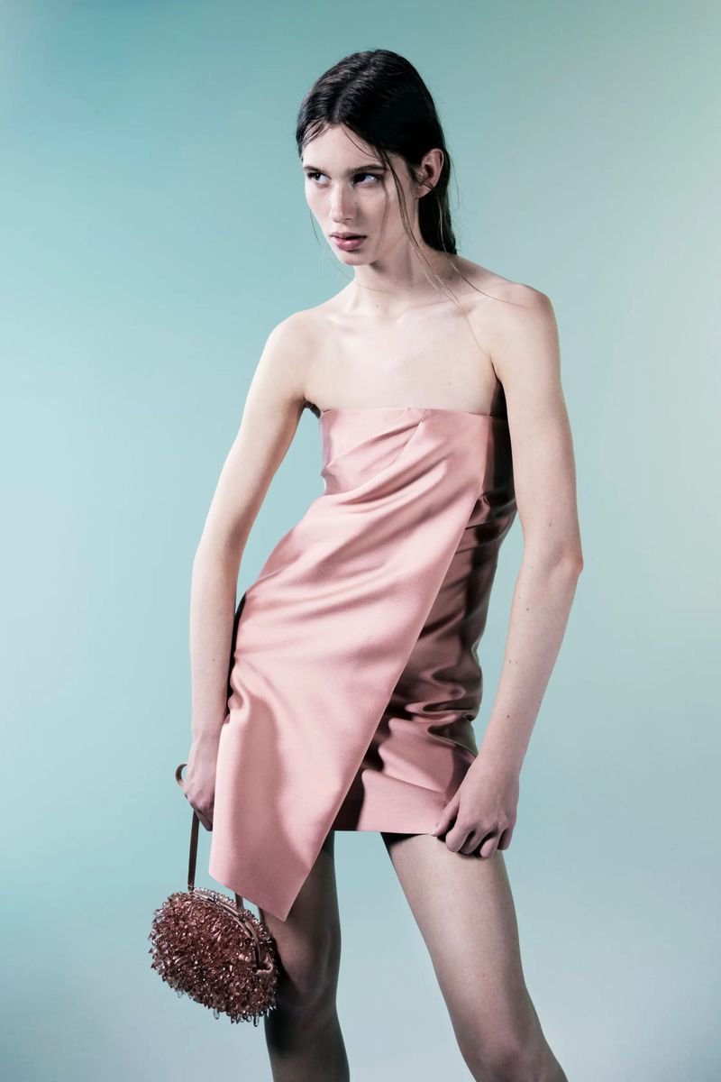 Elegant woman in a pink strapless dress and with beaded bag strikes a pose in a studio with a teal-to-grey gradient background.