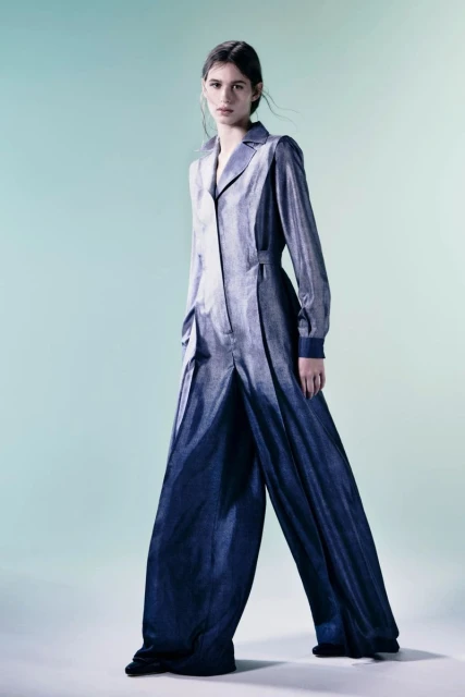 Fashion shot of a woman in a blue metallic wide-legged jumpsuit posing in front of a light blue backdrop in a studio environment.