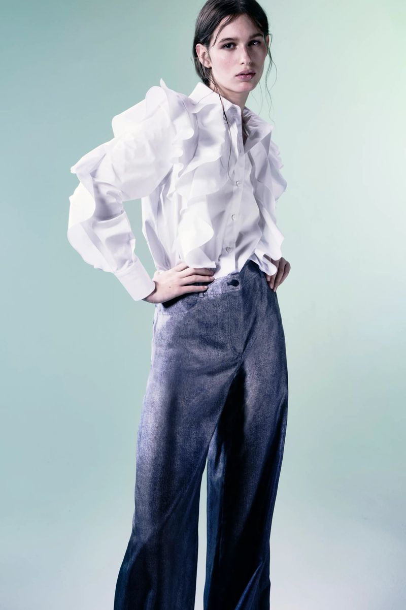 Full shot of a young adult woman in a white ruffle-sleeved shirt and wide leg, blue-gray trousers standing in a photo studio.