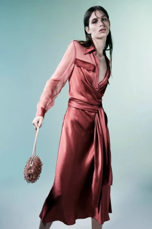A woman in a copper wrap dress and holding a beaded purse poses in front of a graduated aqua background in a fashion studio shot.