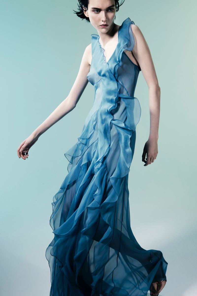 A female model poses in a full-length shot, wearing a flowing blue gown with ruffled detailing, against a gradient blue backdrop in studio.