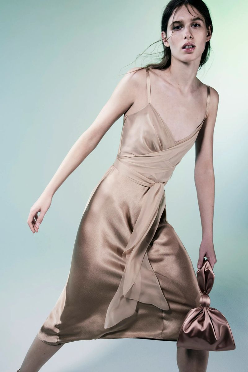 Fashion model in champagne-colored slip dress, strikes pose holding matching satin clutch in studio with blended blue-green background.