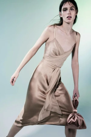 Fashion model in champagne-colored slip dress, strikes pose holding matching satin clutch in studio with blended blue-green background.