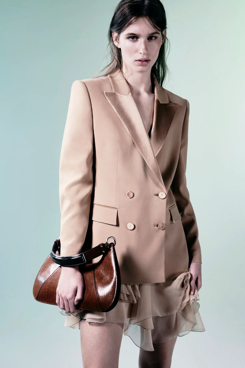 Model wearing a tailored beige double-breasted blazer and sheer ruffled skirt while holding a brown leather handbag in a studio.