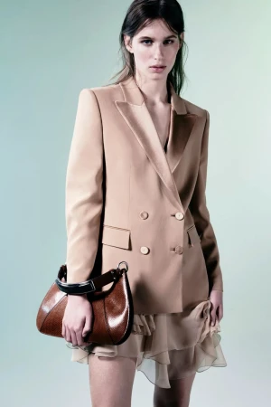 Model wearing a tailored beige double-breasted blazer and sheer ruffled skirt while holding a brown leather handbag in a studio.