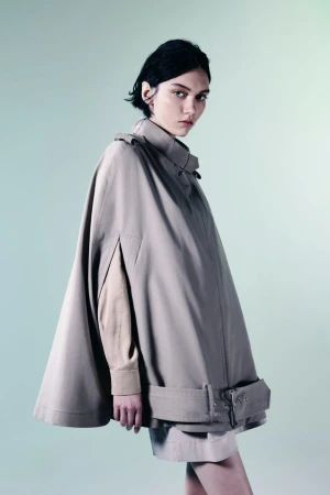 Model wearing a structured beige trench cape with high collar and arm slits against a pale mint gradient studio backdrop.