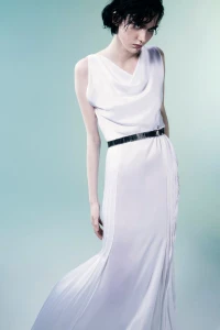 Model posing in a draped sleeveless white floor-length gown with a thin black belt against a pale cyan gradient studio backdrop.