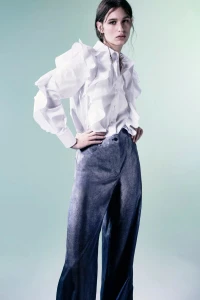 Model posing with hands on hips in a white ruffled button-up shirt and wide-leg grey trousers against a soft gradient backdrop.