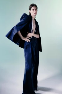 Model posing in a navy blue satin suit with wide-leg trousers and a draped jacket against a soft pale green studio seamless backdrop.