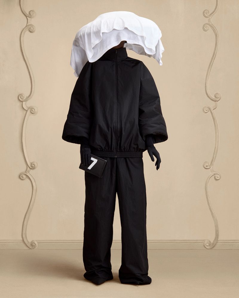 Person in black outfit with white head covering poses against decorative beige wall. Fashion forward style.