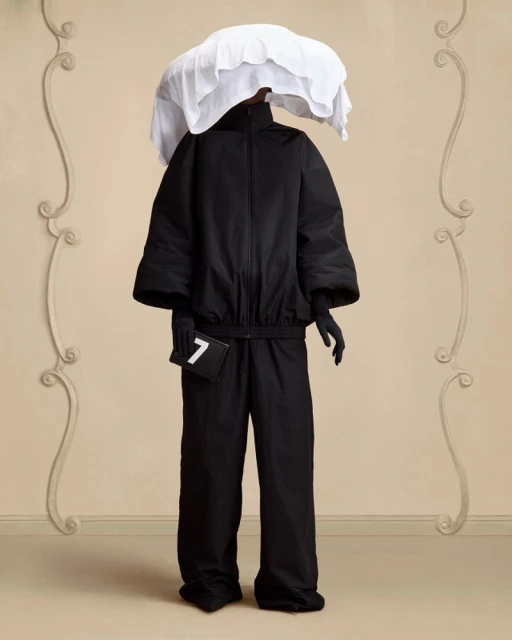 Person in black outfit with white head covering poses against decorative beige wall. Fashion forward style.