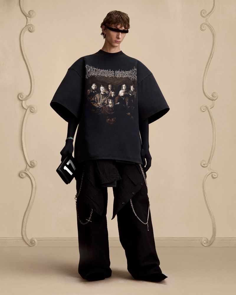 A slender man poses in an oversized black graphic tee, layered pants with chains, and a small black box purse, set against a beige wall.