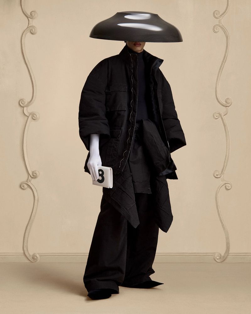 Person poses in avant-garde black padded coat, wide-leg pants, white gloves, and oversized hat with a neutral studio backdrop.