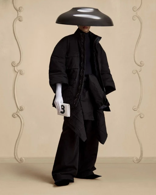 Person poses in avant-garde black padded coat, wide-leg pants, white gloves, and oversized hat with a neutral studio backdrop.