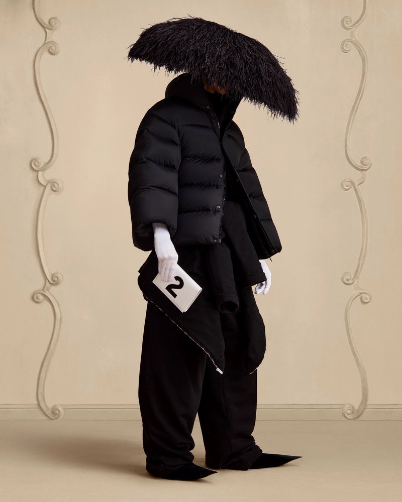 Fashion-forward figure with an ostrich-feathered hat, black puffer jacket, wide trousers, pointed boots, and white-gloved hand holding a tile with the number 2.