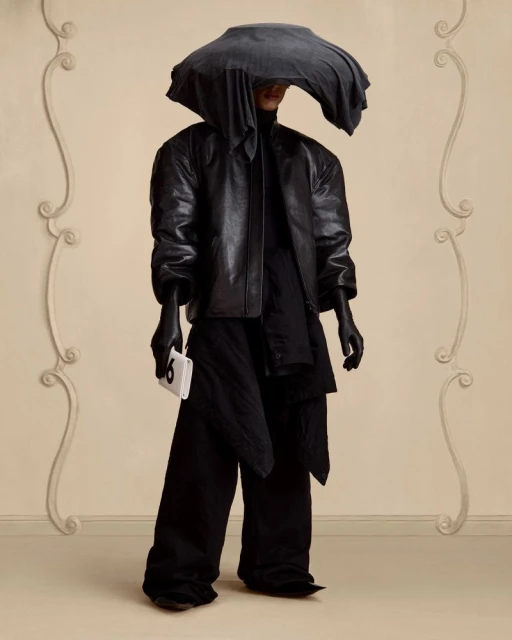 A person wearing a black leather jacket, loose black pants, gloves, and a grey head covering holds a white object against a neutral backdrop.
