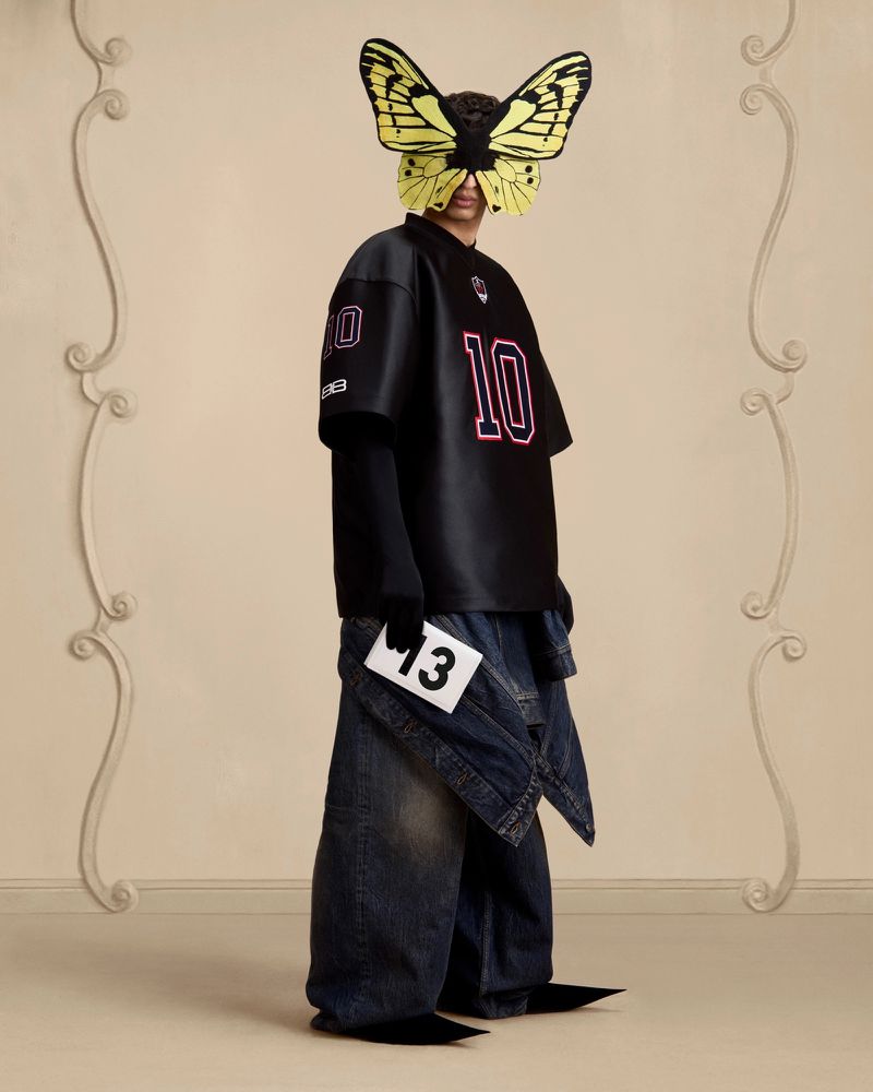 Avant-garde fashion shot: Person wearing a black jersey and denim, face obscured by butterfly mask, holding a card with '13'.