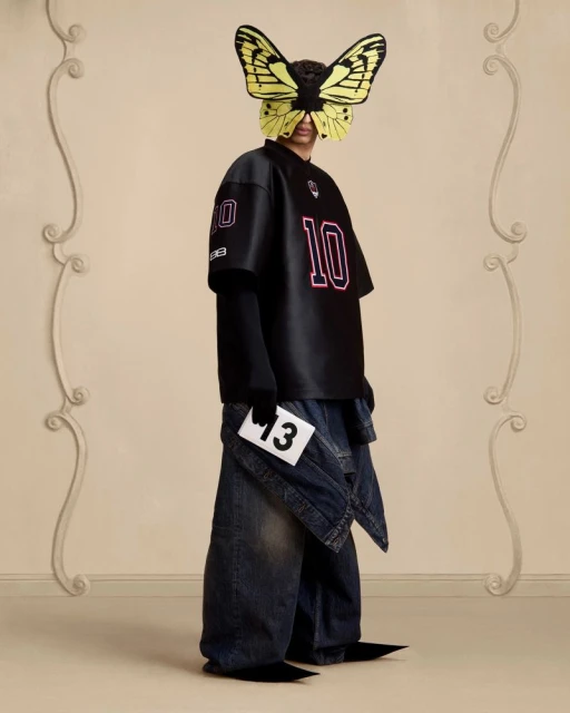 Avant-garde fashion shot: Person wearing a black jersey and denim, face obscured by butterfly mask, holding a card with '13'.