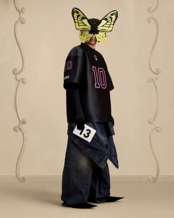 Avant-garde fashion shot: Person wearing a black jersey and denim, face obscured by butterfly mask, holding a card with '13'.