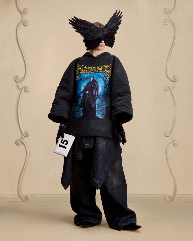 Fashion model wearing a black hoodie with a graphic print, wide denim pants, a feather mask, and holding a white number card against a beige background.