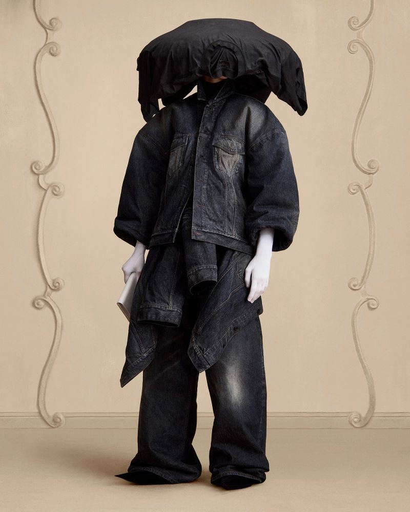 Person dressed in an oversized black denim outfit with a large black head covering stands in front of a beige wall with white moulding.