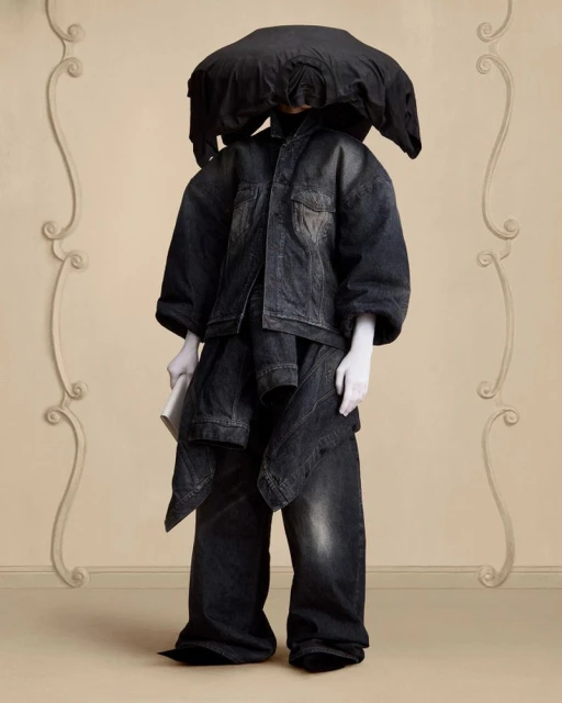 Person dressed in an oversized black denim outfit with a large black head covering stands in front of a beige wall with white moulding.
