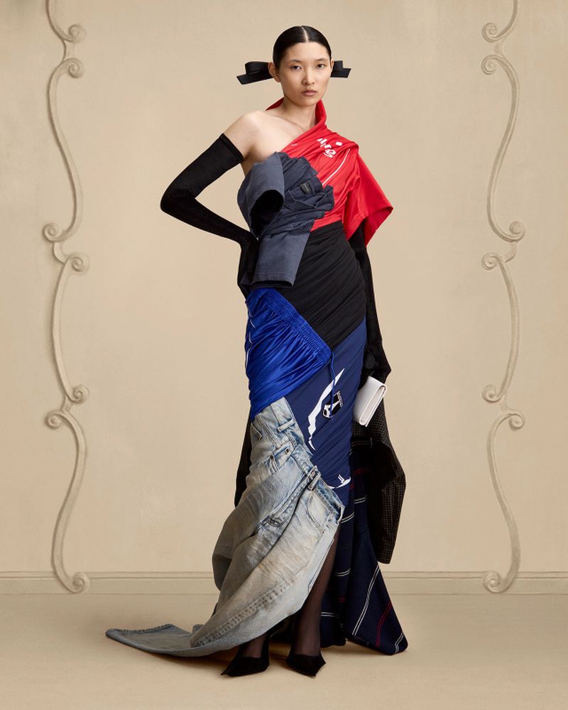 Asian woman in a deconstructed high-fashion gown made of denim, silk, and cotton stands in a beige studio with ornamental wall detail.