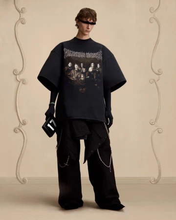 Model posing in an oversized black graphic t-shirt, wide-leg trousers, and eye covering against a beige wall with decorative moldings.