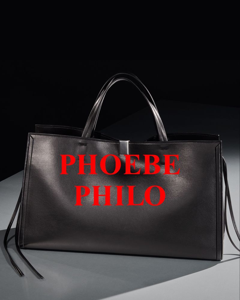 A black leather handbag with two handles stands on a gray surface against a dark background, showcasing the brand name in red text.