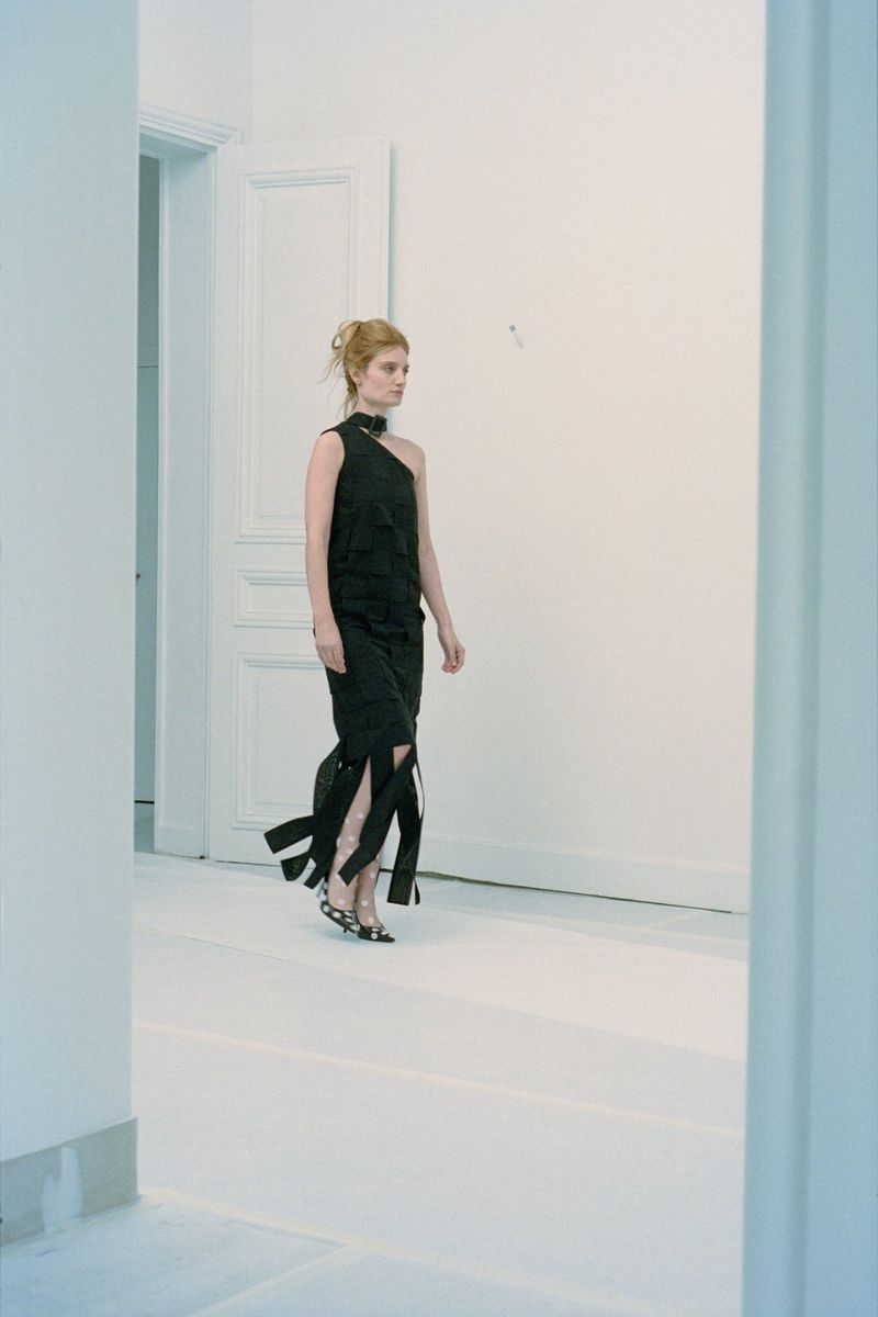 A woman in a dark, one-shoulder dress walks in a light-filled, minimalist interior with white walls and floors, embodying contemporary fashion.