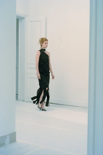 A woman in a dark, one-shoulder dress walks in a light-filled, minimalist interior with white walls and floors, embodying contemporary fashion.