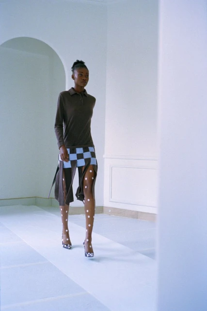 A woman models a brown long-sleeved polo shirt and checkerboard skirt with patterned tights in a minimalist white indoor space.