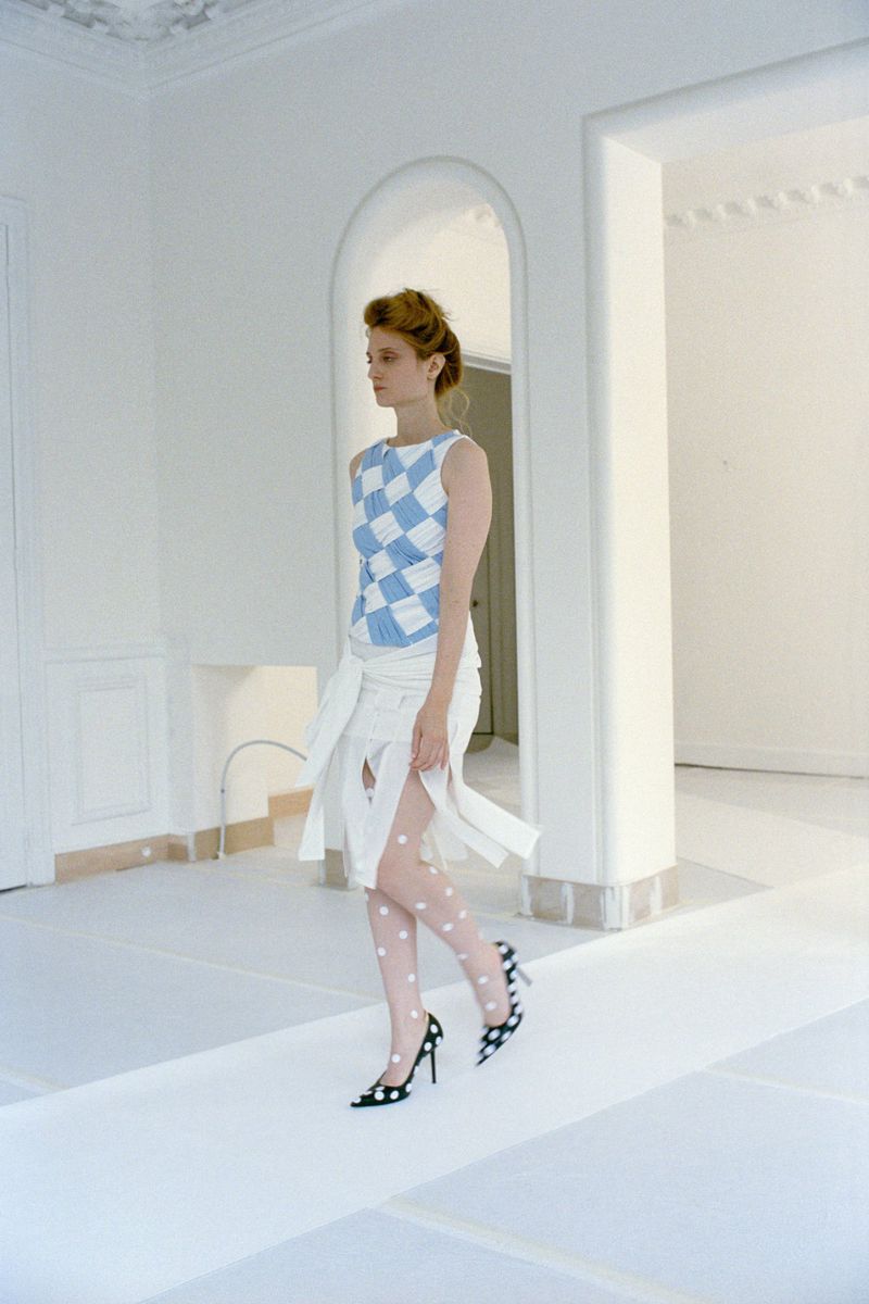 Model walking in a blue and white checkered top and white fringed skirt through an all-white room with an arched doorway.