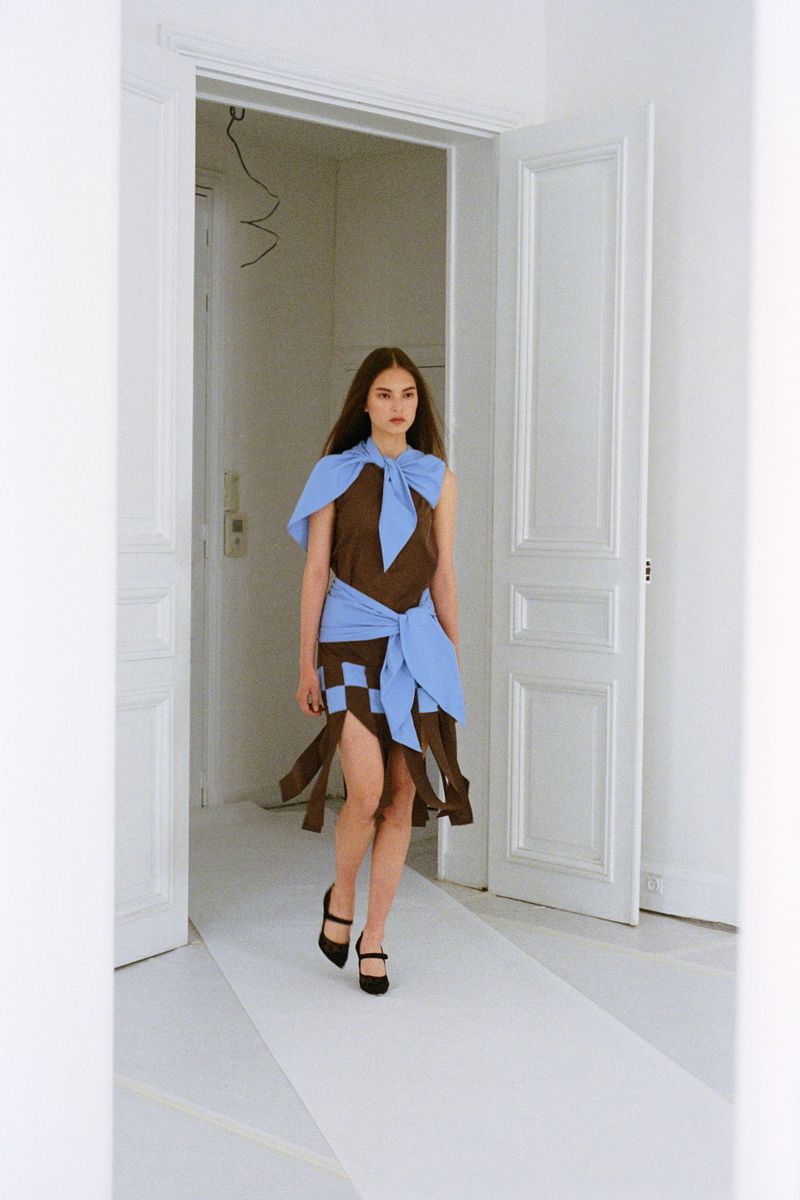Woman in brown and blue patchwork dress and black heels walks down a white catwalk framed by white doors and walls, indoor fashion shot.