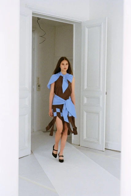 Woman in brown and blue patchwork dress and black heels walks down a white catwalk framed by white doors and walls, indoor fashion shot.