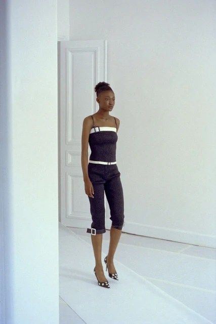 A slender woman with dark skin and her hair up is shown in a fashion pose in a strappy top and capri pants, with a bright white background.