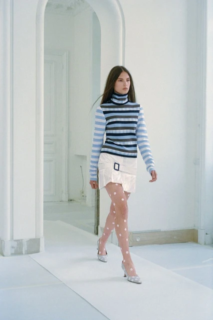 A woman strides confidently in a striped turtleneck, white skirt, and polka dot stockings against a bright white architectural backdrop.