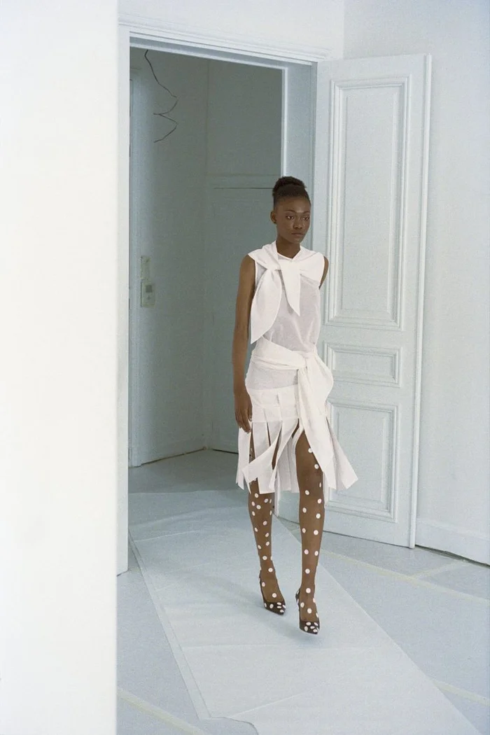 Model walking in a white sleeveless top and skirt with polka-dot tights in a white interior
