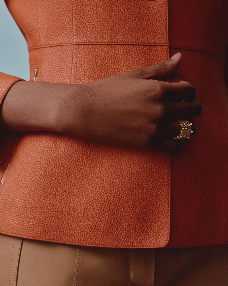 Close-up studio shot of a hand wearing a gold ring with pearls resting against a textured orange leather jacket and tan pants.