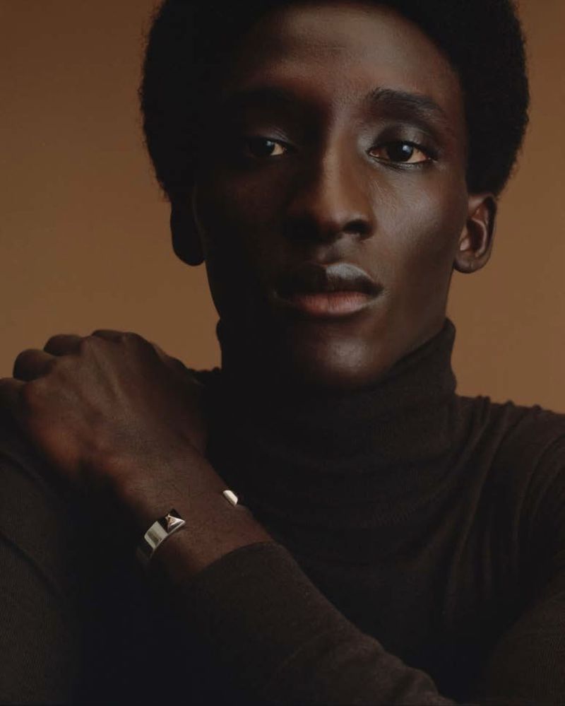 Portrait of a Black man with a dark turtleneck sweater and a silver bracelet against a brown background in a studio setting.