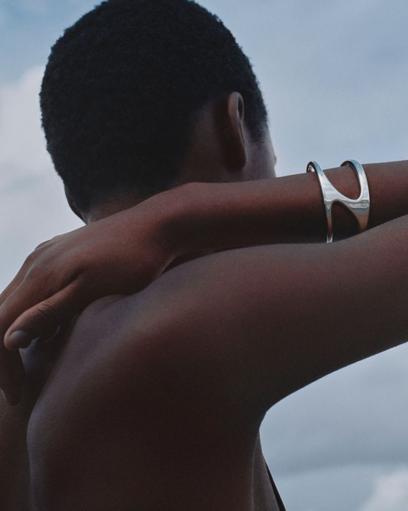 Close up of a person wearing a silver cuff bracelet against a blue and grey sky, with their arms crossed in front of their torso.