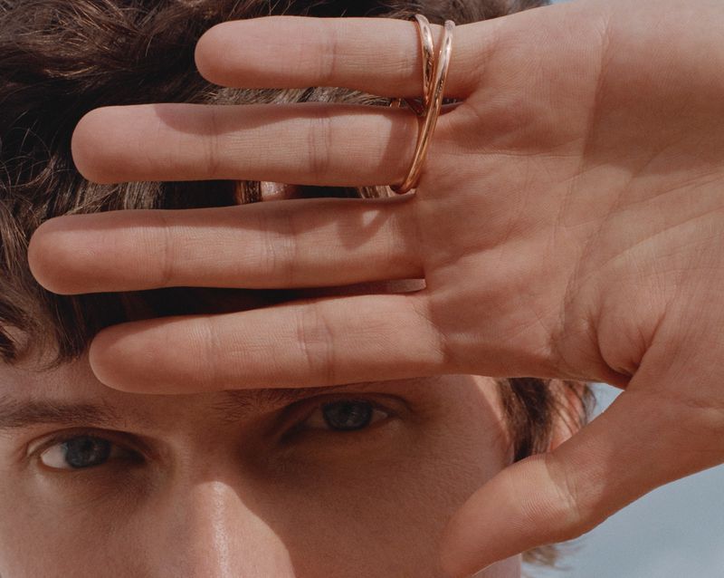 Close-up of a fair-skinned adult male with blue eyes and brown hair, his face partially obscured by his raised hand and a metal ring.
