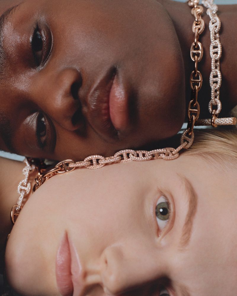 Close-up portrait of two diverse women with fair and dark skin, each wearing a chain-link necklace with diamond accents. Beauty shot.