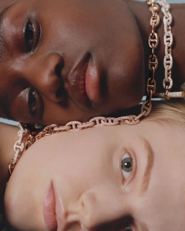 Close-up portrait of two diverse women with fair and dark skin, each wearing a chain-link necklace with diamond accents. Beauty shot.
