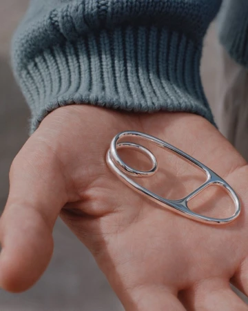 Close-up of a silver metal decorative safety pin in an adult's hand with a light blue sweater sleeve in soft focus as a background element.