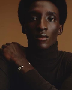 Close-up portrait of a Black man wearing a dark turtleneck and silver cuff bracelet against a warm neutral background.