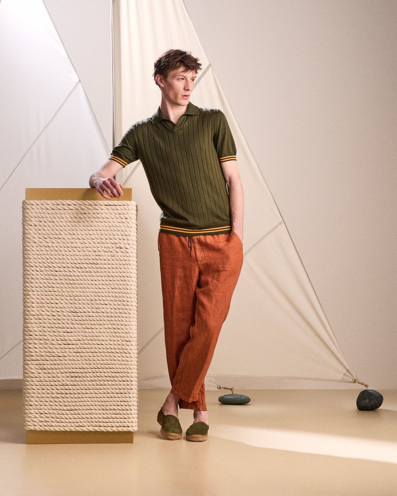 Male model in olive polo and orange pants leaning on rope pillar in a fashion setting, showcasing modern menswear and studio photography.